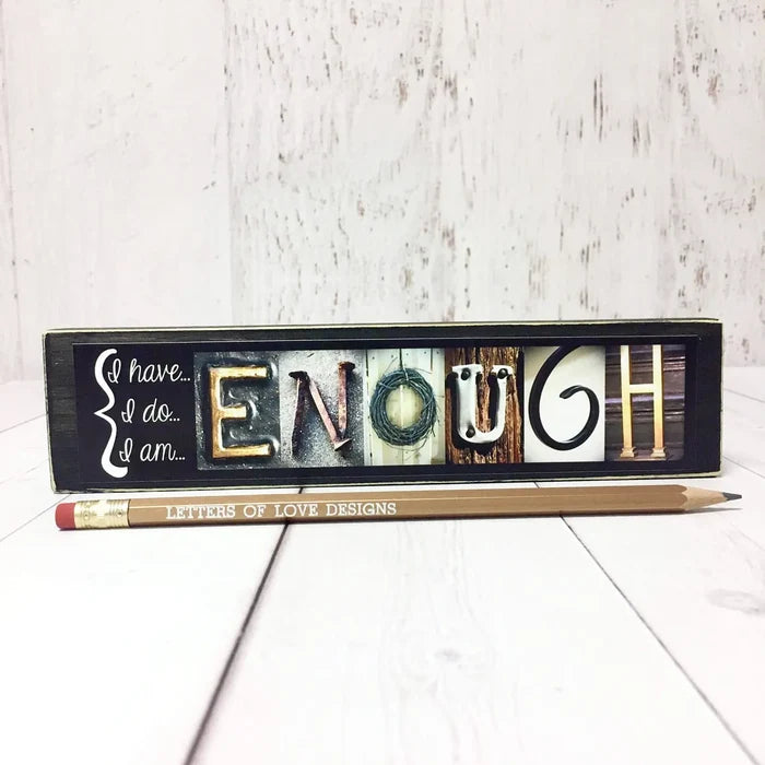 Decorative sign with letter tiles spelling 'ENOUGH' on a wooden background, with a pencil in front.