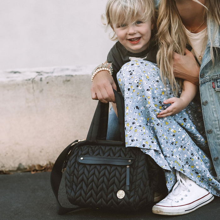 Mom holding mini structured quilted diaper bag by the handles while out and about with her toddler.